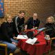A photograph of four people sitting at a round table, which has microphones and other podcast equipment on it